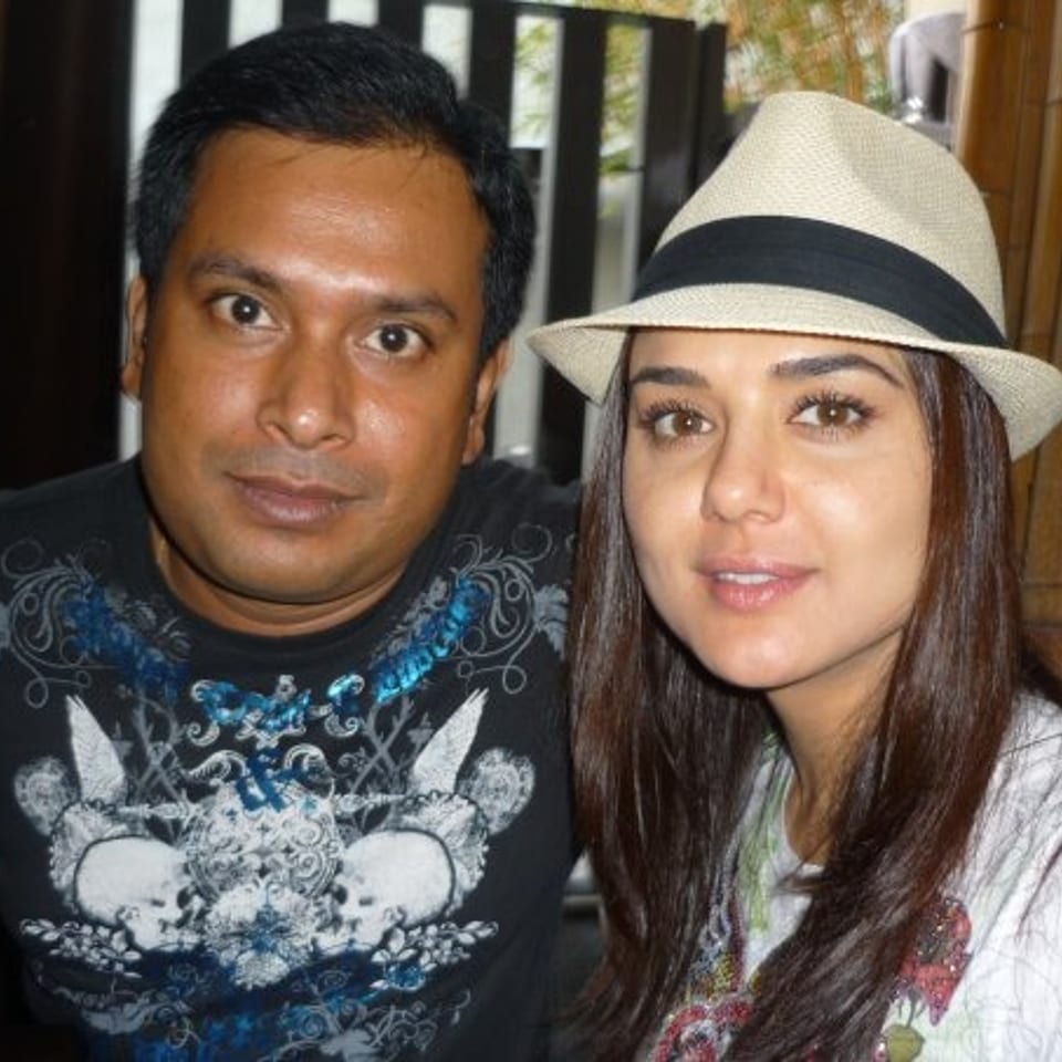 Two people pose together indoors, with the woman wearing a white fedora and the man in a graphic black T-shirt.