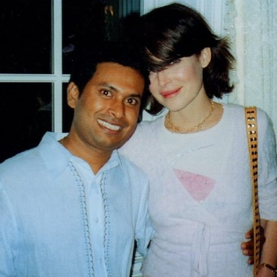 Two smiling people pose together indoors, with a window behind them and one person carrying a studded shoulder bag.