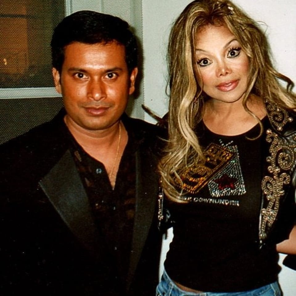 Man and woman pose together indoors, wearing black outfits, with the woman in a decorative jacket and jeans.