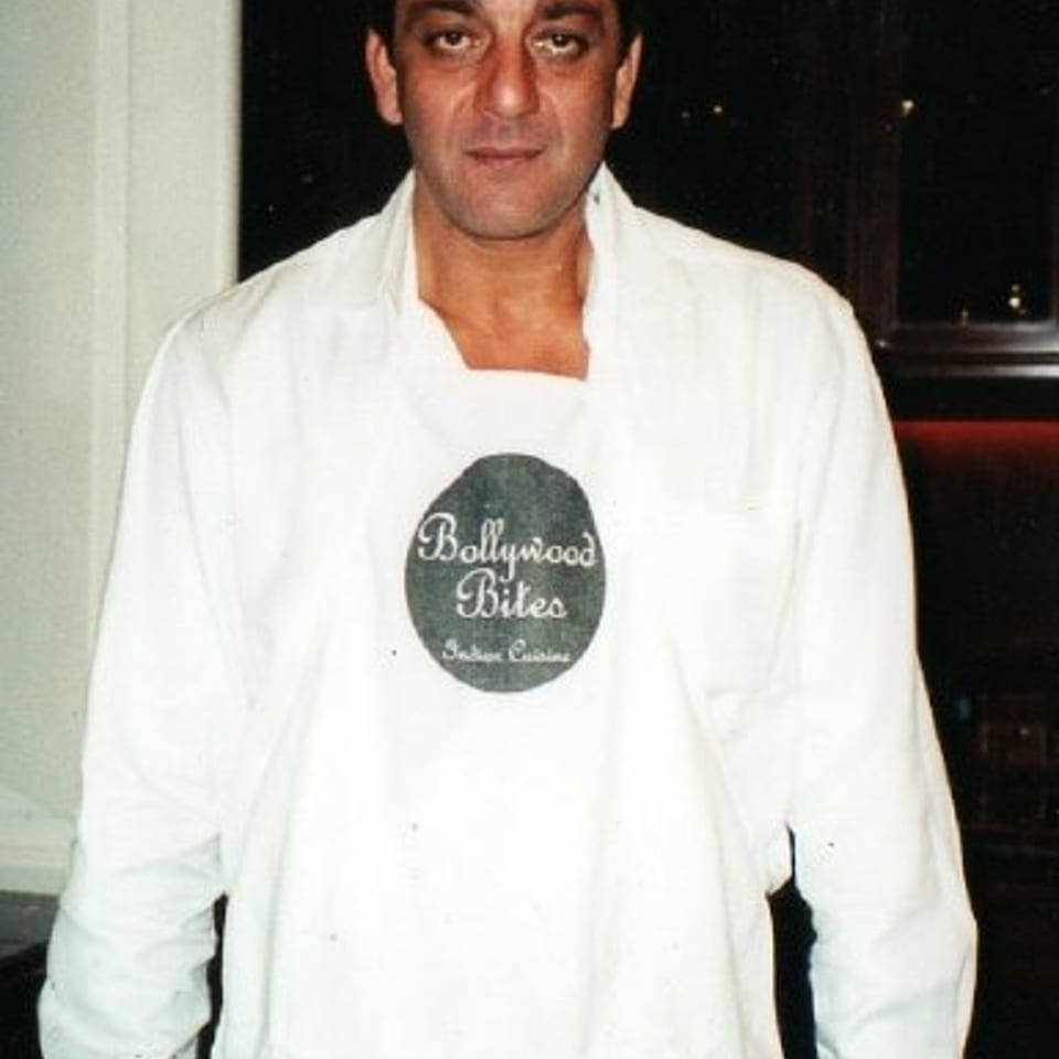 Man wearing a white chef’s coat and Bollywood Bites apron, standing indoors.