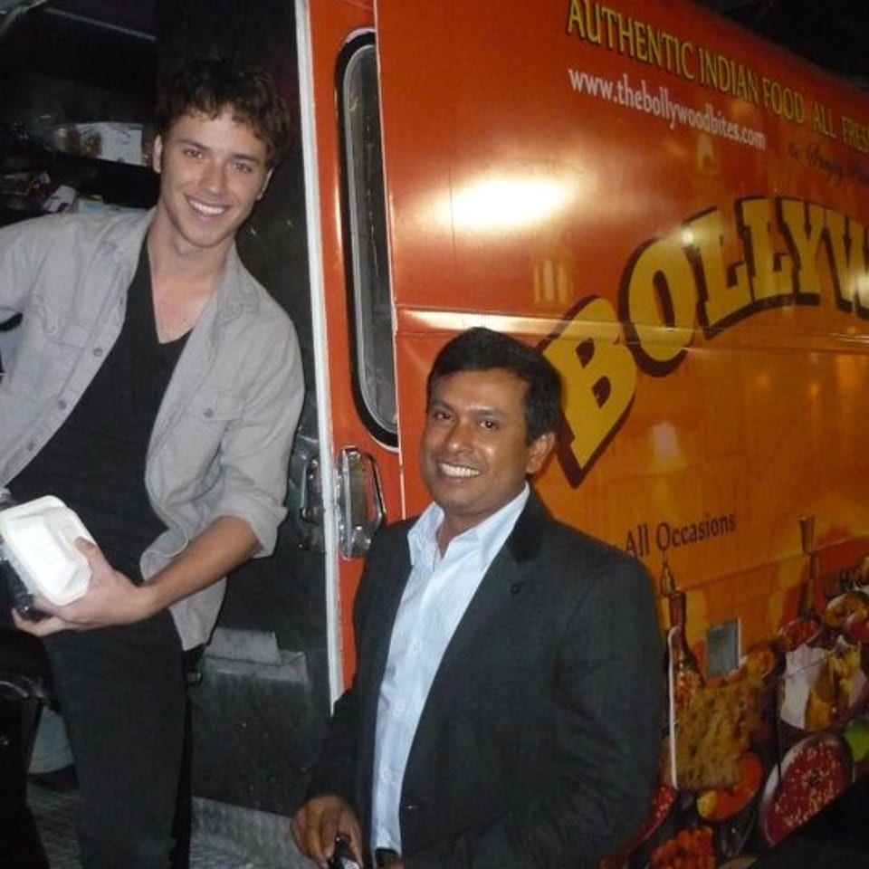 Two smiling men stand beside an orange Bollywood-themed Indian food truck, with one holding a takeout container.