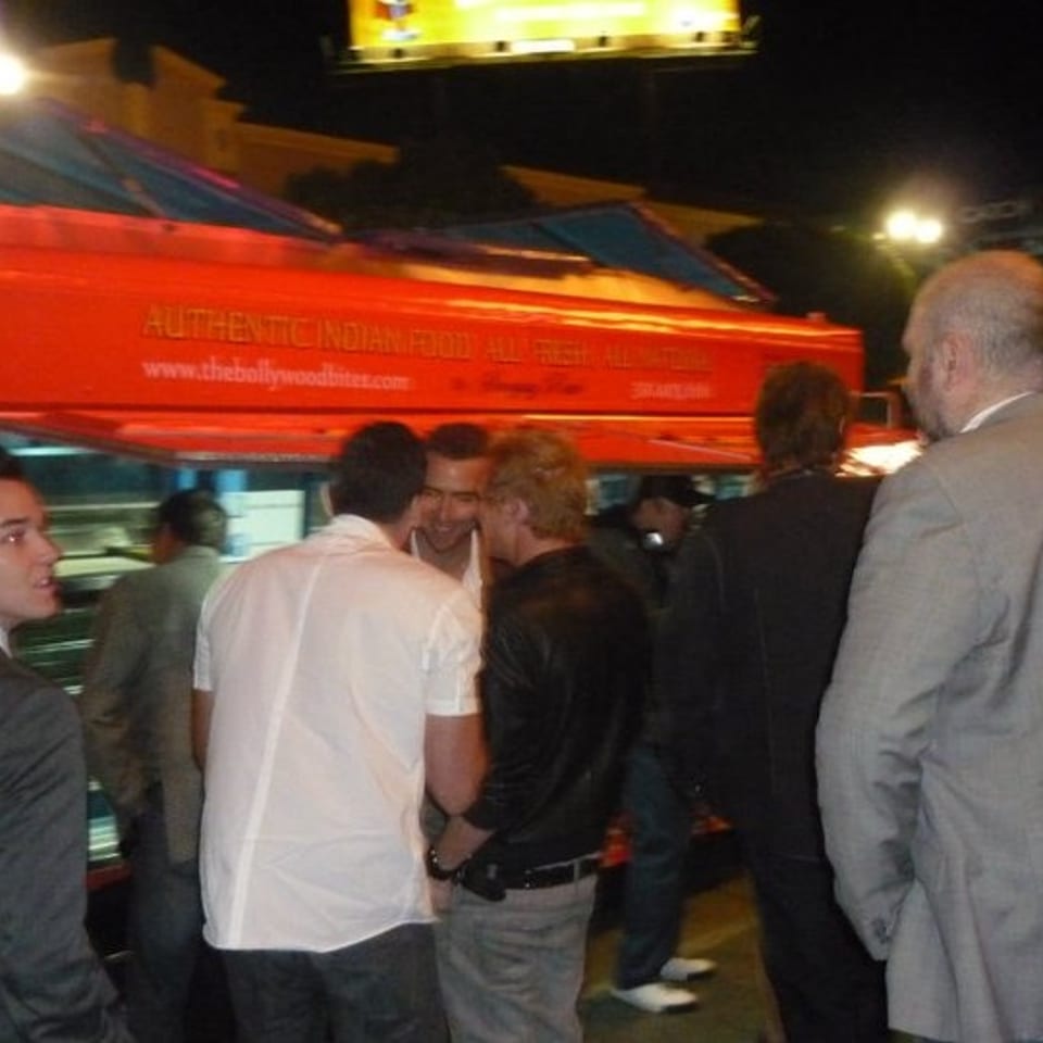 People gather around a bright red Indian food truck at night, with illuminated signs and city lights overhead.