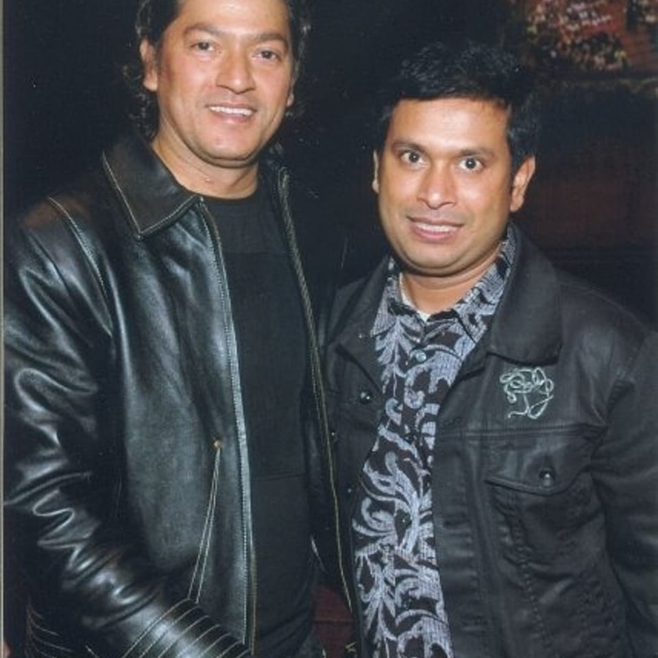Two men pose together indoors, wearing dark leather jackets; one wears a patterned shirt in a dimly lit setting.