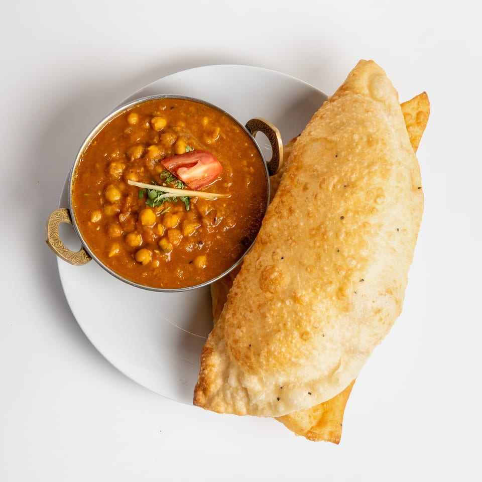 A bowl of chickpea curry with a slice of tomato and ginger, next to a large fried puri.