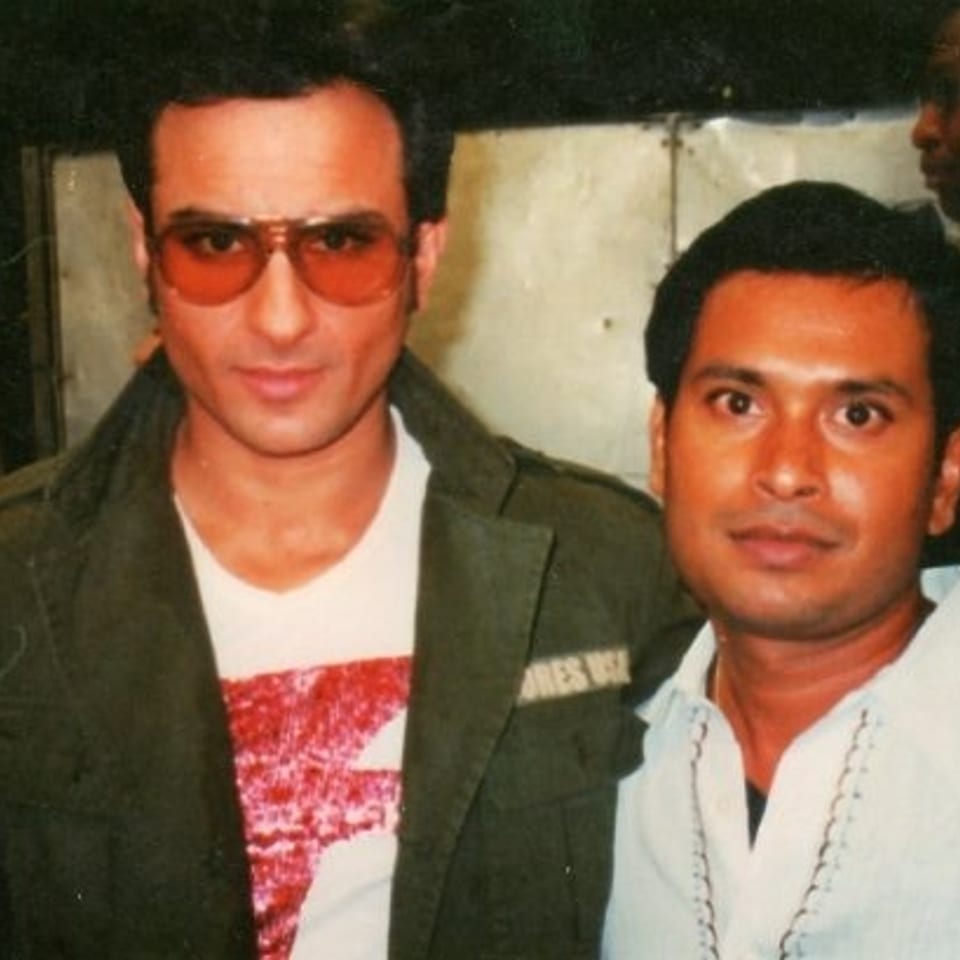 Two men pose together indoors, one wearing sunglasses and a green jacket, amid several people in the background.