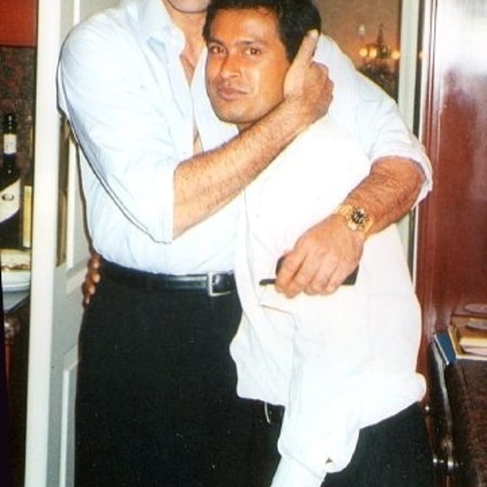 Two men in white shirts embrace indoors near a doorway, smiling at the camera.