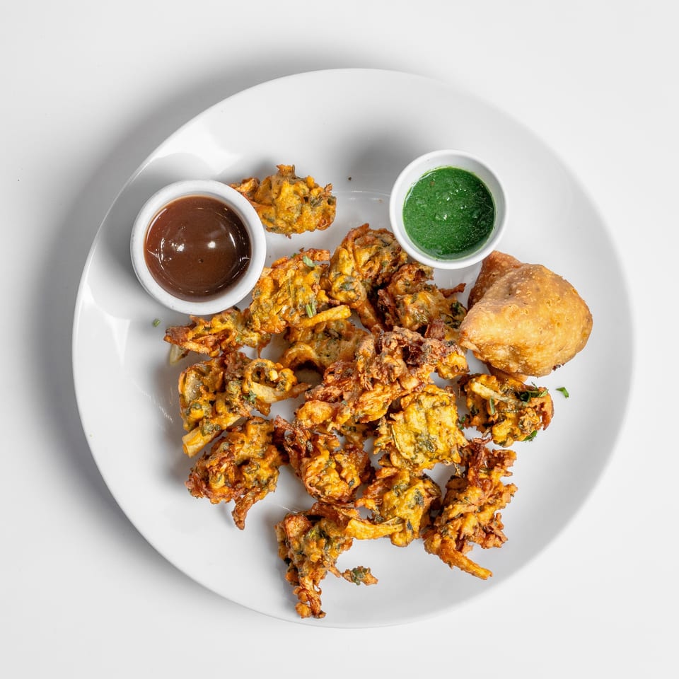A white plate with pakoras, a samosa, and two dipping sauces, one brown and one green.