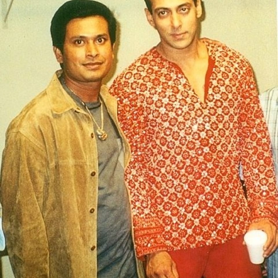 Two men pose indoors, one wearing a tan jacket and the other a red patterned shirt while holding a white cup.