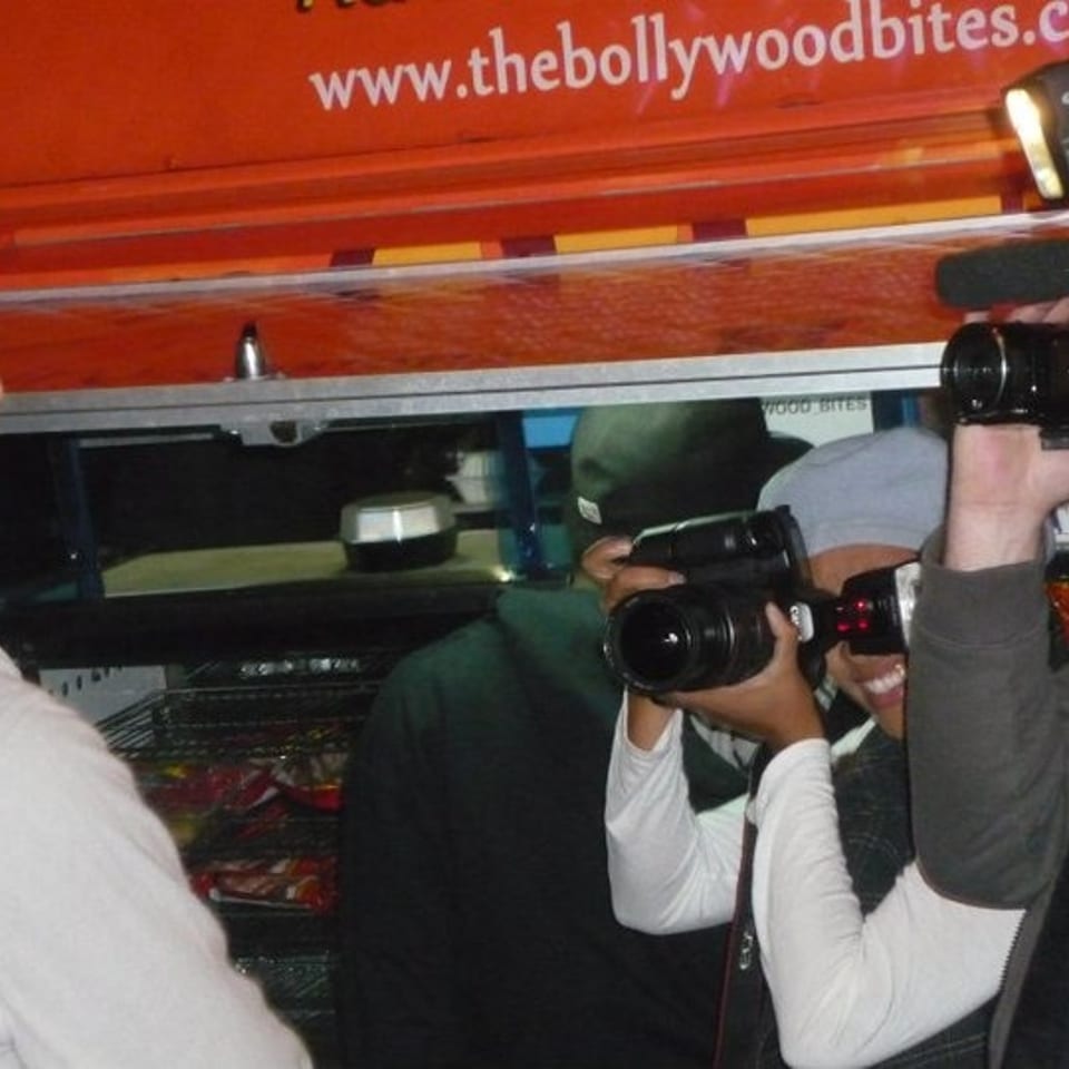 Photographers film beside a red Bollywood Bites food truck displaying its website.
