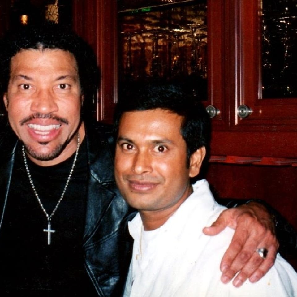 Two men pose together inside a dimly lit restaurant, one wearing a cross necklace and the other dressed in white.