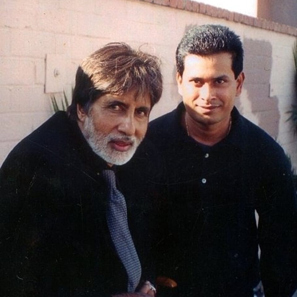 Two men in black clothing pose together outdoors beside a light-colored brick wall.