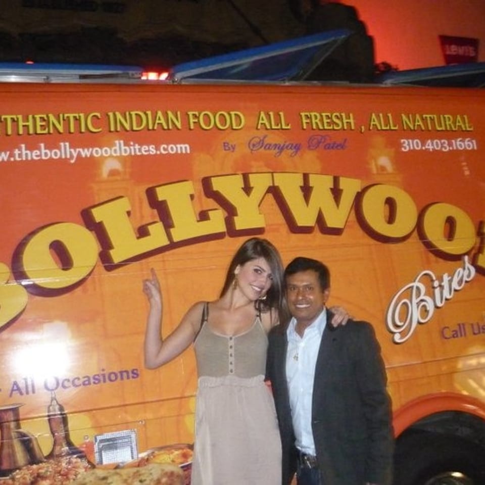 Orange Bollywood Bites food truck advertising authentic Indian cuisine, with two people posing beside it.