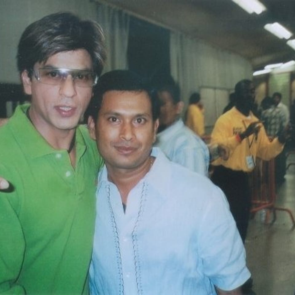 Two men pose together in a crowded indoor venue, with people and stalls visible in the background.