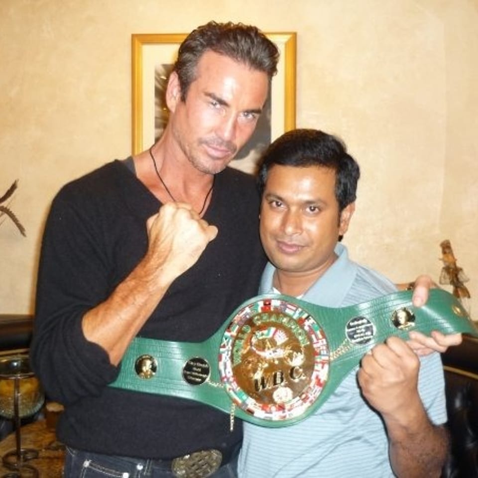 Two men pose indoors, holding a green championship wrestling belt with a large central medallion.