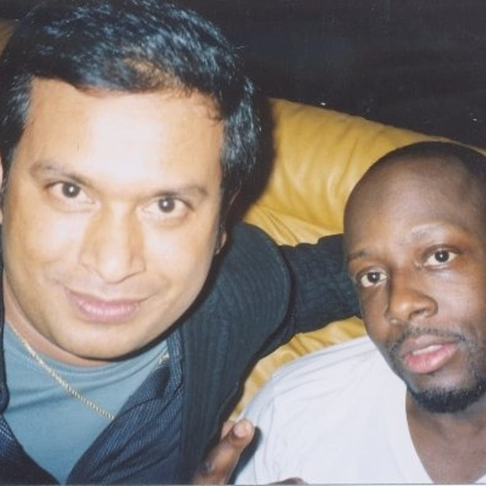 Two men pose together on a yellow leather couch, smiling toward the camera in a dimly lit indoor setting.