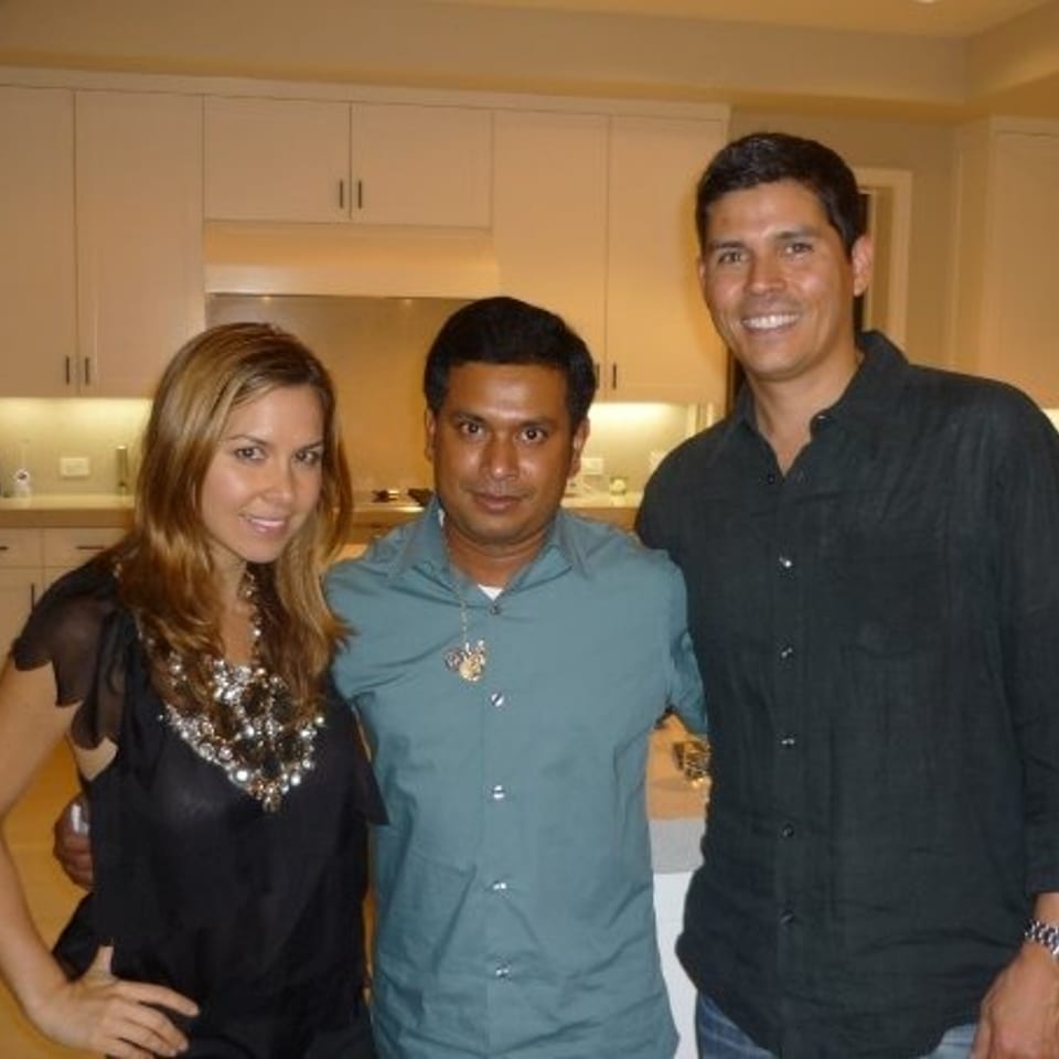 Three smiling adults pose together in a warmly lit modern kitchen with white cabinets and countertops.