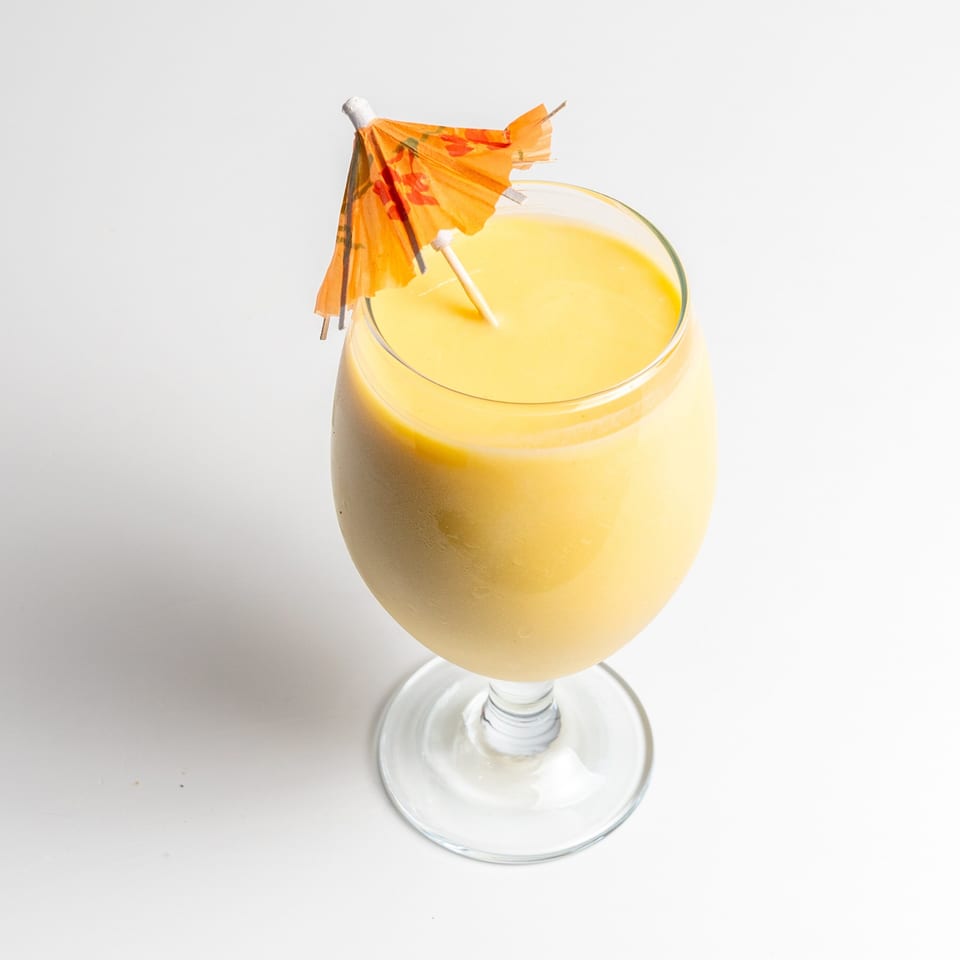 A creamy yellow mango lassi in a wine glass with a small orange paper umbrella.