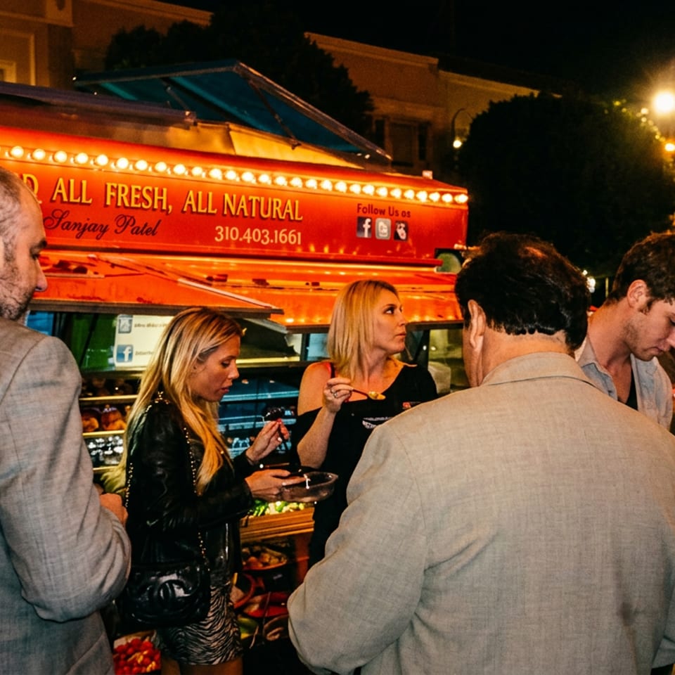 People gather around a brightly lit red food truck at night, some eating, others signing.