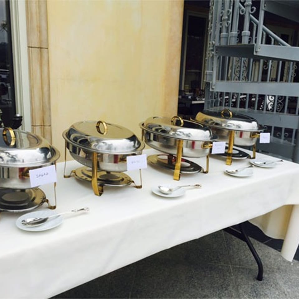 Outdoor buffet setup with four silver chafing dishes on a white-covered table, one labeled "SALAD".